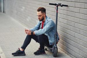 beau jeune homme en vêtements décontractés assis sur un scooter électrique à l'extérieur pendant la journée ensoleillée photo