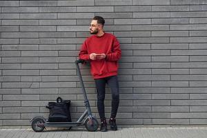 avec sac noir. beau jeune homme en vêtements décontractés debout avec un scooter électrique à l'extérieur pendant la journée ensoleillée photo