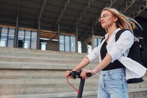 à proximité d'un bâtiment commercial. Belle blonde en vêtements décontractés équitation schooter électrique à l'extérieur à la journée ensoleillée photo