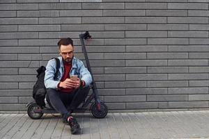 beau jeune homme en vêtements décontractés assis sur un scooter électrique à l'extérieur pendant la journée ensoleillée photo