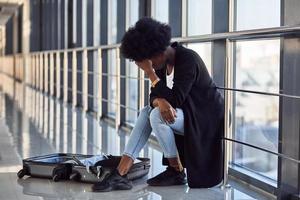 triste et déprimée jeune passagère afro-américaine en tenue décontractée assise à l'aéroport avec bagages photo