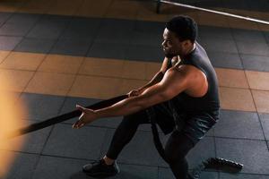 un homme afro-américain fort en vêtements sportifs a une journée d'entraînement dans la salle de sport. faire du crossfit en utilisant des cordes photo