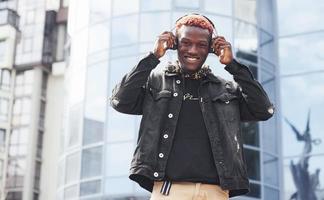 écouter de la musique dans des écouteurs. jeune homme afro-américain en veste noire à l'extérieur dans la ville debout contre un bâtiment d'affaires moderne photo