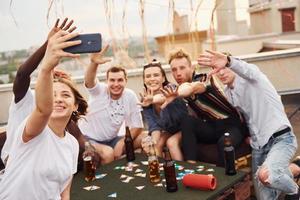 fille faisant une photo quand les gens jouent au jeu de cartes. un groupe de jeunes en vêtements décontractés fait la fête ensemble sur le toit pendant la journée