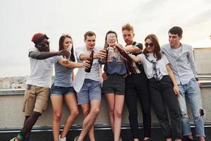 debout avec de l'alcool au bord du toit. un groupe de jeunes en vêtements décontractés fait la fête ensemble pendant la journée photo
