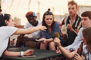 assis à table et jouant au jeu de cartes. un groupe de jeunes en vêtements décontractés fait la fête ensemble sur le toit pendant la journée photo