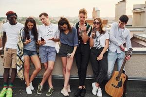 debout avec des téléphones et de l'alcool dans les mains. un groupe de jeunes en vêtements décontractés fait la fête ensemble sur le toit pendant la journée photo