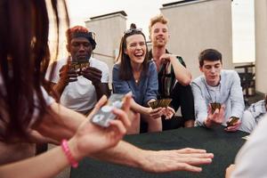 assis à table et jouant au jeu de cartes. un groupe de jeunes en vêtements décontractés fait la fête ensemble sur le toit pendant la journée photo
