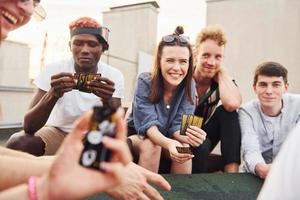 assis à table et jouant au jeu de cartes. un groupe de jeunes en vêtements décontractés fait la fête ensemble sur le toit pendant la journée photo