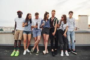 debout avec de l'alcool au bord du toit. un groupe de jeunes en vêtements décontractés fait la fête ensemble pendant la journée photo