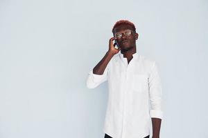 jeune homme afro-américain en vêtements formels blancs parlant au téléphone contre le mur à l'intérieur photo