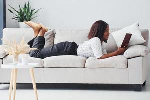 femme afro-américaine positive en chemise blanche allongée sur un canapé avec un bloc-notes dans les mains photo