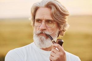 portrait d'un homme senior élégant aux cheveux gris et à la barbe qui se tient à l'extérieur sur le terrain à la journée ensoleillée et qui fume photo