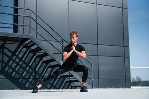 échauffement près du bâtiment noir. jeune homme sportif en chemise noire et pantalon à l'extérieur photo