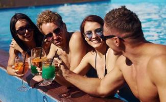 parler et boire. un groupe de jeunes gens heureux s'amuse dans la piscine pendant la journée photo