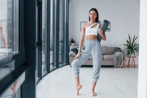 debout et tient le tapis. jeune femme avec une forme de corps mince en vêtements de sport a une journée de remise en forme à l'intérieur à la maison photo