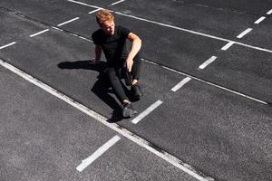 le coureur fatigué est assis sur la piste et fait une pause. jeune homme sportif en chemise noire et pantalon à l'extérieur pendant la journée photo