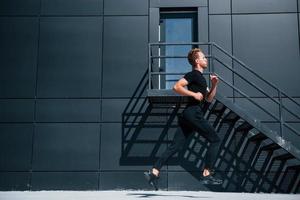 courir près du bâtiment noir. jeune homme sportif en chemise noire et pantalon à l'extérieur pendant la journée photo