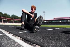 le coureur fatigué est assis sur la piste et fait une pause. jeune homme sportif en chemise noire et pantalon à l'extérieur pendant la journée photo