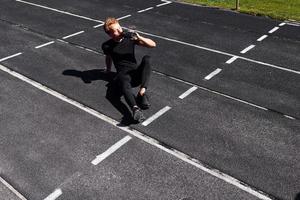 le coureur fatigué est assis sur la piste et fait une pause. jeune homme sportif en chemise noire et pantalon à l'extérieur pendant la journée photo