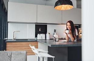 avec une tasse de café frais. jeune belle brune en vêtements décontractés à l'intérieur dans la cuisine pendant la journée photo