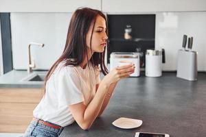 jeune belle brune en tenue décontractée est assise dans la cuisine avec une boisson fraîche le matin photo