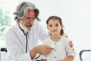 Médecin senior aux cheveux gris et à la barbe en blouse blanche mesurant la fréquence cardiaque d'une petite fille par stéthoscope à l'intérieur d'une clinique photo