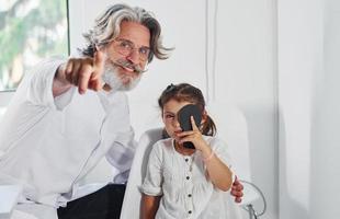 ophtalmologiste masculin senior aux cheveux gris et à la barbe en blouse blanche est à l'intérieur dans une clinique testant la vue d'une petite fille photo