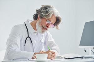 écrit par table d'ordinateur. médecin senior aux cheveux gris et à la barbe en blouse blanche est à l'intérieur de la clinique photo