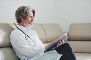 médecin senior aux cheveux gris et à la barbe en blouse blanche est à l'intérieur de la clinique est assis sur un canapé avec un bloc-notes dans les mains photo