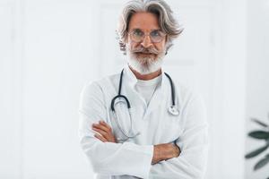 portrait d'un médecin senior aux cheveux gris et à la barbe en blouse blanche debout à l'intérieur d'une clinique photo