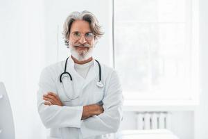 portrait d'un médecin senior aux cheveux gris et à la barbe en blouse blanche debout à l'intérieur d'une clinique photo