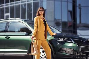 posant pour un appareil photo. jeune femme à la mode en manteau de couleur bordeaux pendant la journée avec sa voiture photo