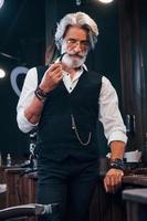 debout et fumer dans un salon de coiffure. homme âgé moderne et élégant aux cheveux gris et à la barbe est à l'intérieur photo