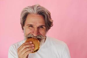 mange un délicieux burger. homme âgé moderne et élégant aux cheveux gris et à la barbe est à l'intérieur photo
