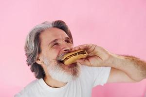 mange un délicieux burger. homme âgé moderne et élégant aux cheveux gris et à la barbe est à l'intérieur photo