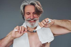 à l'aide de dentifrice. homme âgé moderne et élégant aux cheveux gris et à la barbe est à l'intérieur photo