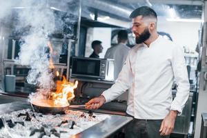 poêle à frire est en feu. chef en uniforme blanc cuisinant des aliments à la cuisine. journée bien remplie au travail photo