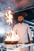 utilise une poêle à frire. chef en uniforme blanc cuisinant des aliments à la cuisine. journée bien remplie au travail photo