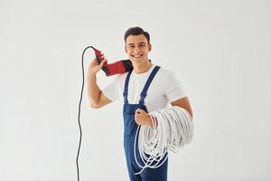 avec perceuse et câbles dans les mains. travailleur masculin en uniforme bleu debout à l'intérieur du studio sur fond blanc photo