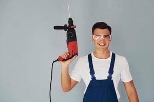 dans des lunettes de protection et avec une perceuse dans les mains. travailleur masculin en uniforme bleu debout à l'intérieur du studio sur fond blanc photo