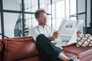 lit le journal. jeune homme caucasien en chemise blanche élégante à l'intérieur à la maison photo