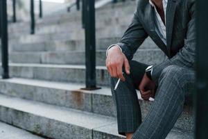 est assis et fume une cigarette. jeune homme d'affaires prospère en tenue de soirée grise est à l'extérieur de la ville photo