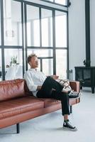 détente sur le canapé. jeune homme caucasien en chemise blanche élégante à l'intérieur à la maison photo