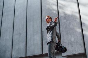 en lunettes de soleil. jeune homme d'affaires prospère en tenue de soirée grise est à l'extérieur de la ville photo