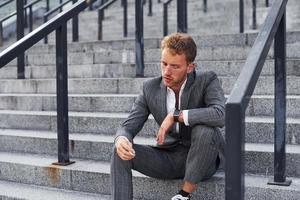 est assis et fume une cigarette. jeune homme d'affaires prospère en tenue de soirée grise est à l'extérieur de la ville photo