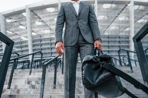 tient le sac dans les mains. jeune homme d'affaires en tenue de soirée grise est à l'extérieur de la ville photo