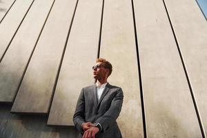 debout contre le mur du bâtiment. jeune homme d'affaires en tenue de soirée grise est à l'extérieur de la ville photo