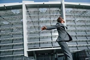 danser, célébrer le succès. jeune homme d'affaires en tenue de soirée grise est à l'extérieur de la ville photo