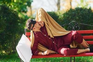 allongé sur un banc dans le parc. jeune homme en pyjama est à l'extérieur dans la rue. se sent somnolent photo